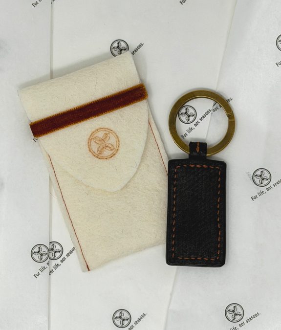Beige wallet with a brown stripe lying beside a brass circular key ring on white branded paper.